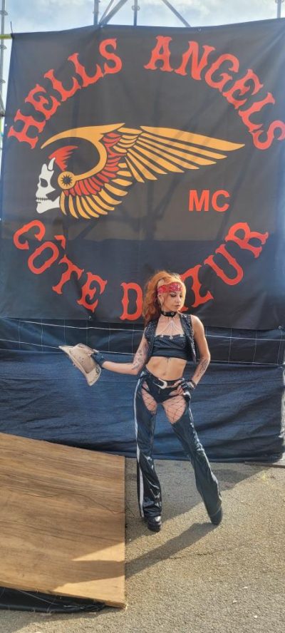 Hell's Week Fréjus 2025 : Stéphanie Molly, performeuse, en tenue cuir noir et chapeau, devant le drapeau Hells Angels Côte d'Azur